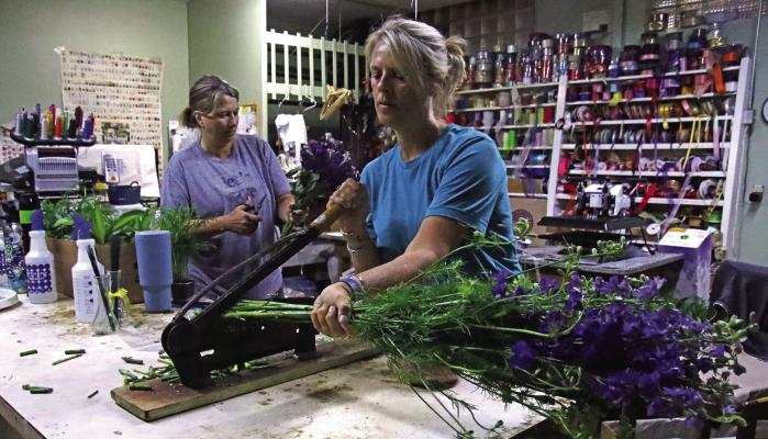 Sharla Long(left) cuts the packaging off of a group of flowers while Jo Lee Ebbers(right), the owner of Blooms, uses a large blade to cut the ends off.